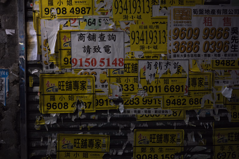A collection of overlapping, colorful advertisements and signs posted on a dark, textured wall, featuring various phone numbers like "9150 5148" and "93419313".
