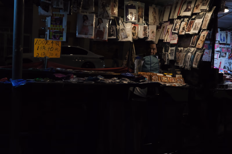A dimly lit street scene with a shopfront displaying various products and a person sitting behind the counter.