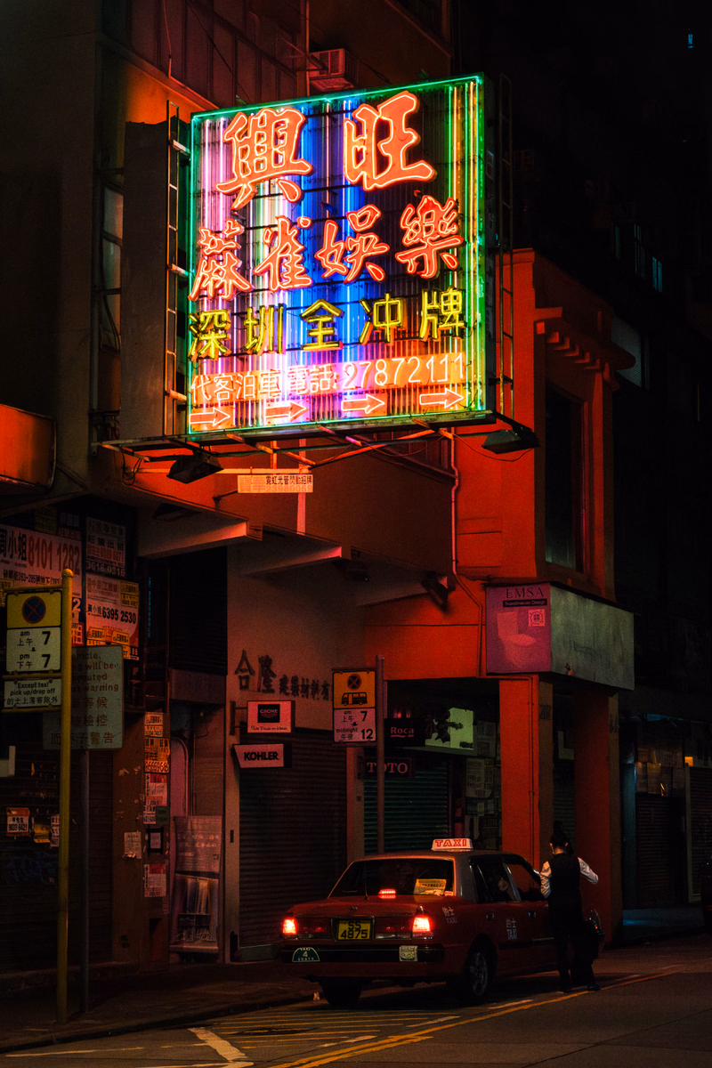A brightly lit neon sign in Chinese characters, which reads "興旺全中牌", illuminates a street scene at night.