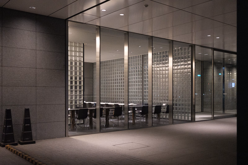 A modern office interior features a glass partition separating a conference room with tables and chairs from a hallway.