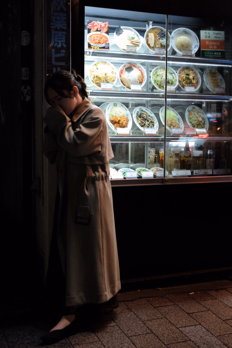 A woman looks at a refrigerated display case menu inside a shop, which features various ramen dishes.