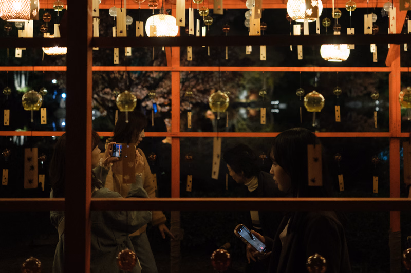 Several people are gathered outdoors at night beneath numerous glowing paper lanterns hanging from a wooden structure.