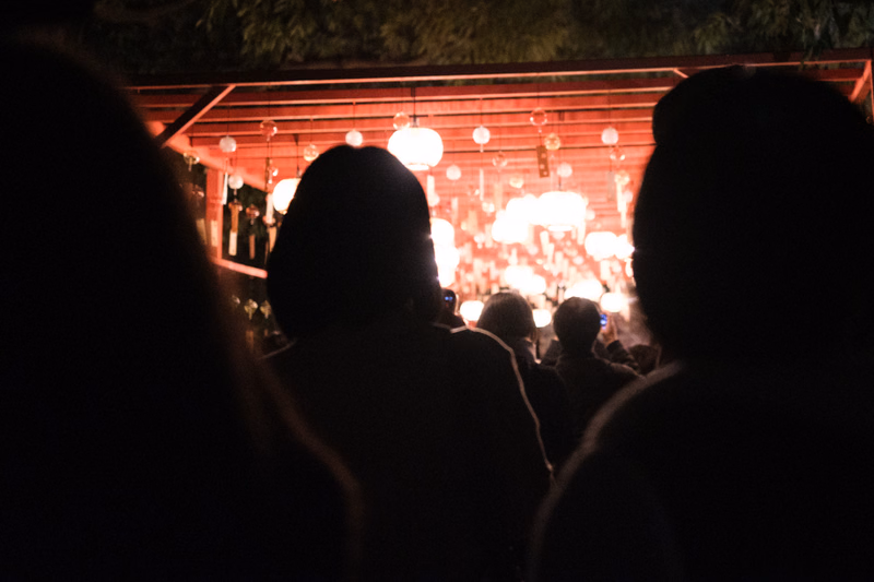 Silhouettes of people attend an outdoor evening gathering illuminated by numerous hanging lights under a wooden structure.