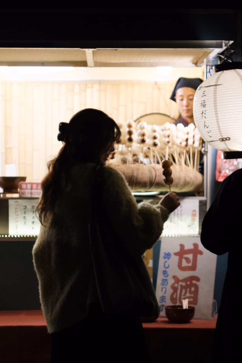 A woman in a light sweater stands inside a shop, holding a small cup, with a white paper lantern displaying "甘酒" visible.