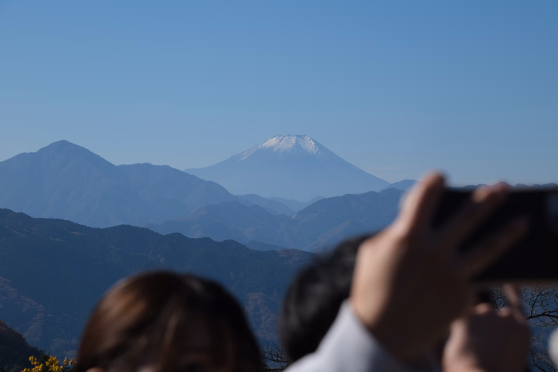 A view of Mount Fuji rising above layers of blue mountains under a clear blue sky, with a person holding a camera in the foreground.
