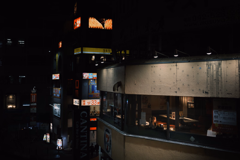 A nighttime view of a commercial area featuring several storefronts, including one with a visible "7-Eleven" logo and another with illuminated signs.