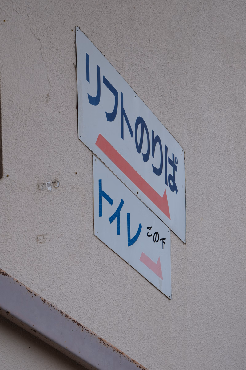 A white sign with Japanese text reads "リフトは トイレの下" which translates to "The lift is under the toilet" with red and blue arrows.