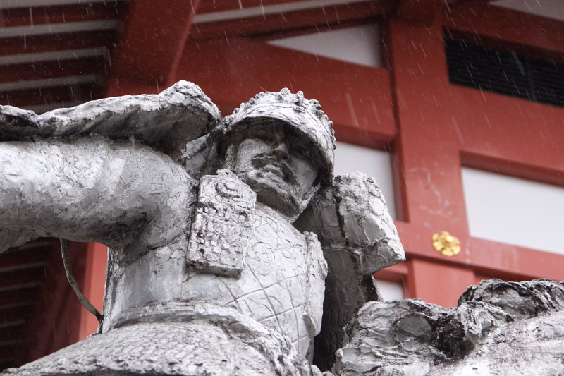 A close-up of a stone statue of a warrior figure being hit by heavy rain in front of a red building.