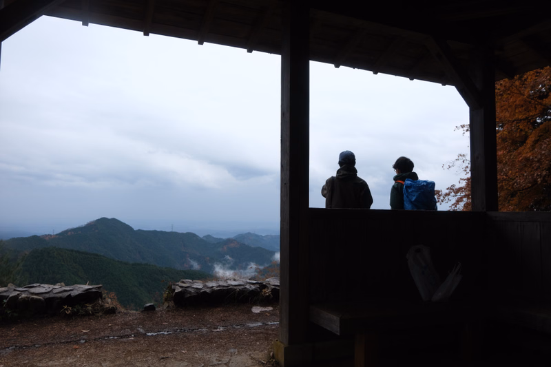 Two hikers stand inside a wooden shelter overlooking a misty mountain landscape under an overcast sky.