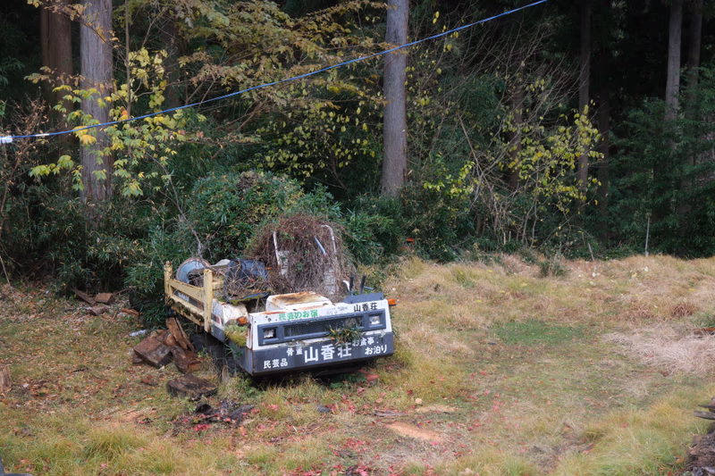 A truck loaded with a large brush pile is parked near a forested area. The truck is labeled with the name 'Itsukaichi' and is situated in a grassy area surrounded by trees.