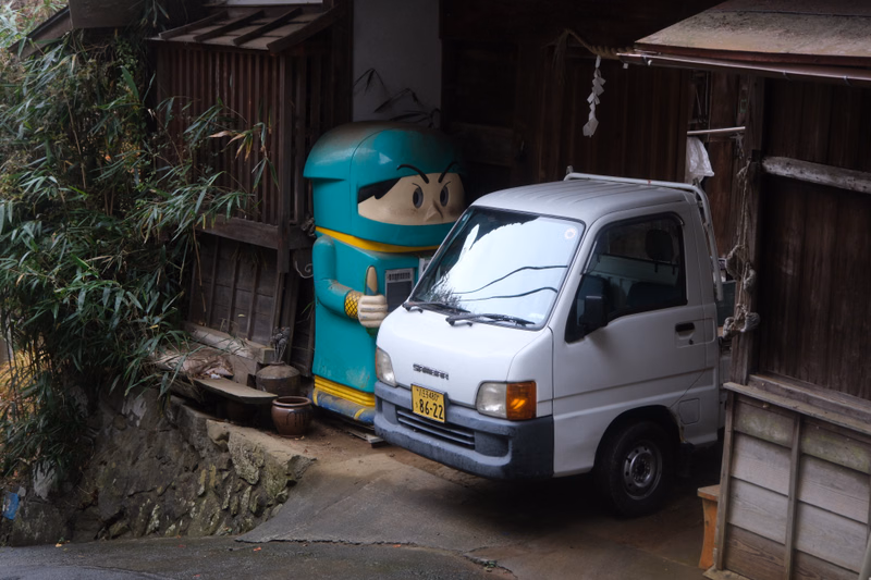 A white van is parked next to a large green and blue character statue outside a wooden house, with bamboo foliage nearby.