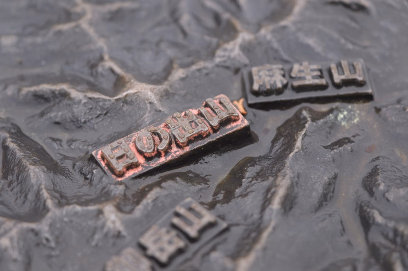 A small, reddish-brown metal tag with engraved Japanese characters, including "麻生山", rests on a dark, textured surface.