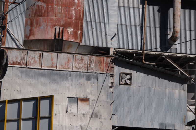 A large industrial building with corrugated metal siding features a prominent red storage tank and various pipes running along the exterior.