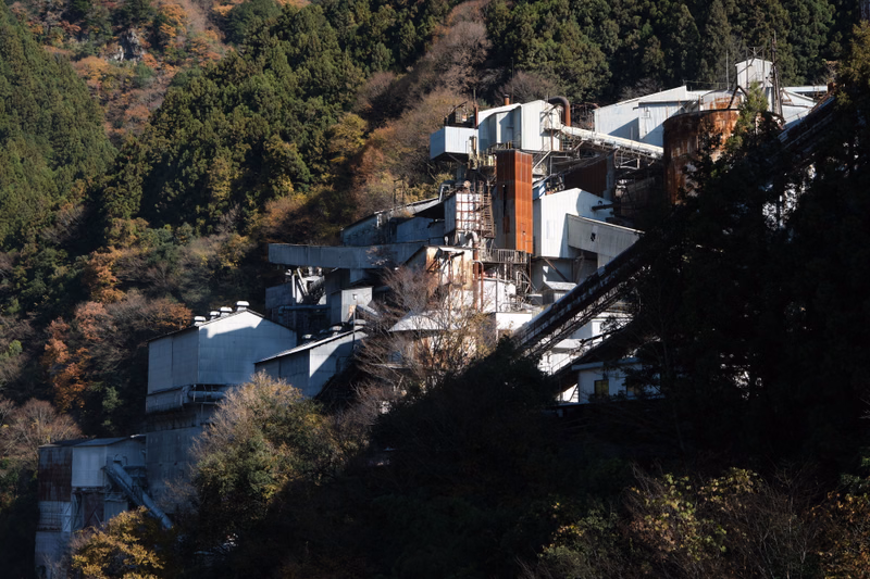 A large, sprawling industrial complex with multiple buildings is nestled into a steep, forested mountainside.