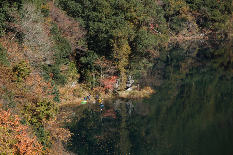 Several small kayaks are visible on the calm, dark water of a lake surrounded by dense, colorful autumn trees.