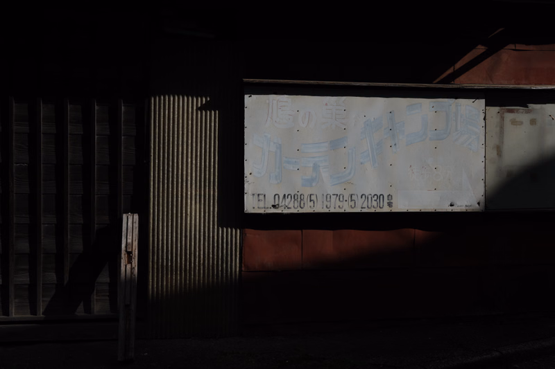 A weathered white sign on a building wall reads "店の名" and "TEL. 04288(5) 1979-52030" in Japanese characters.