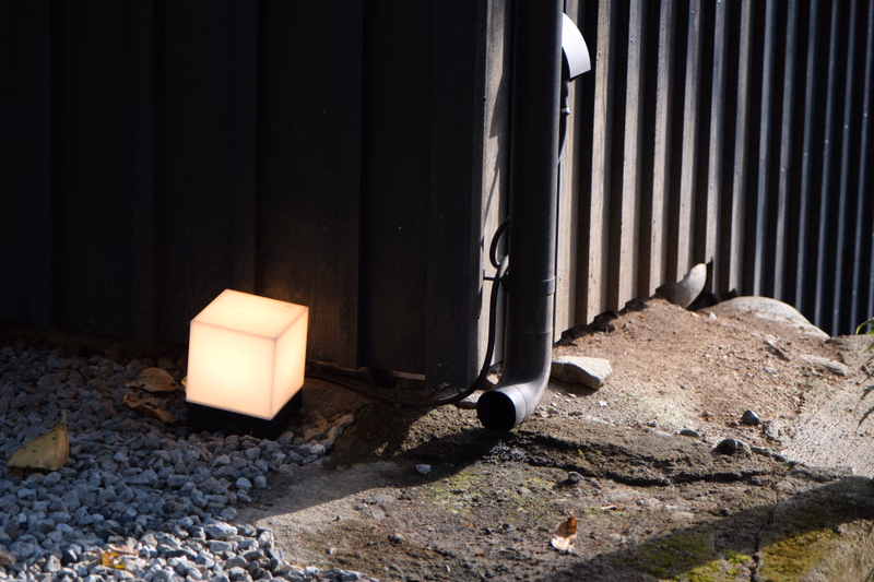 A glowing square lantern sits on gravel next to a dark corrugated metal shed with a vertical vent pipe.