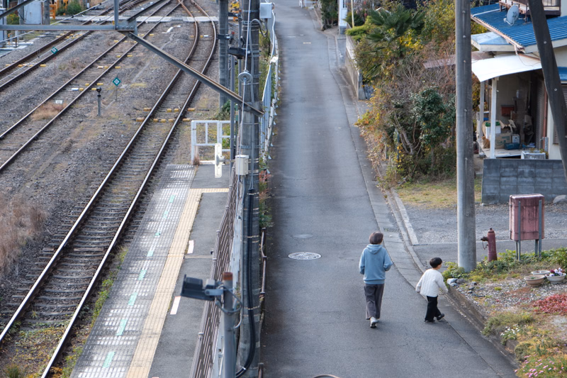 Two people walk down a paved pedestrian path alongside railway tracks next to a residential area.