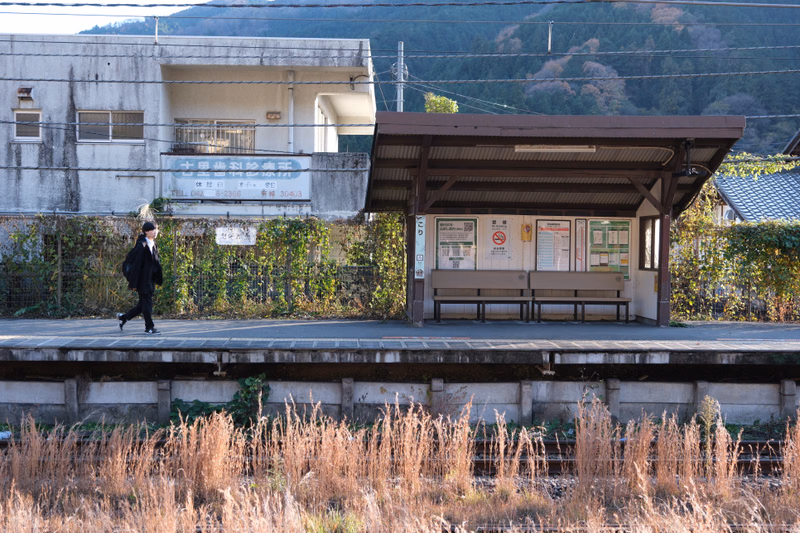 A person walks across the platform of a train station with a covered shelter, next to a light-colored station building.