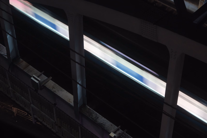 A modern train passes through a covered station platform illuminated by bright, long strip lights.