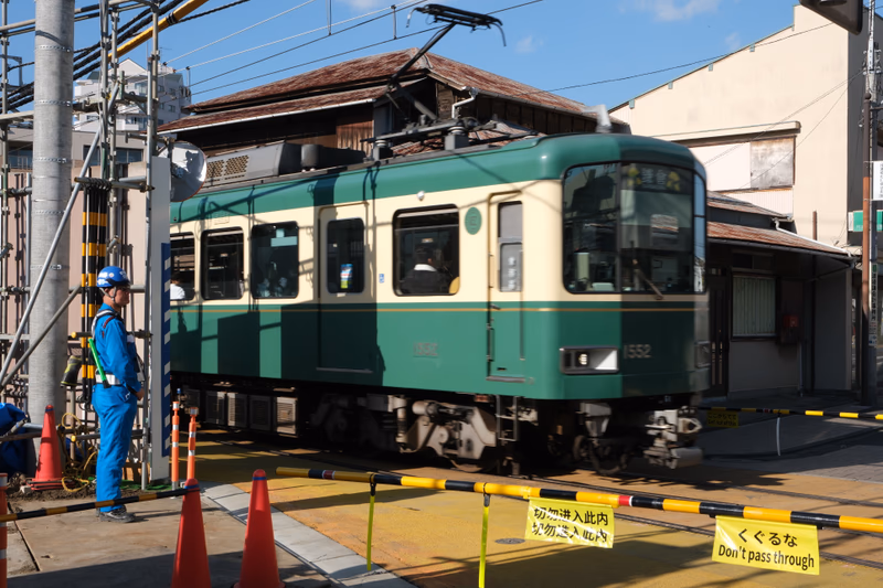 A green and cream-colored train is seen at a railway crossing in Yokohama, Kanagawa, Japan. A person in a blue safety helmet and a blue jumpsuit is standing near the train.