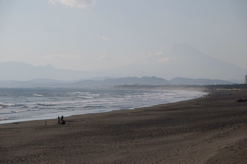 A wide view of a sandy beach meeting the ocean, with a large mountain visible in the hazy background.