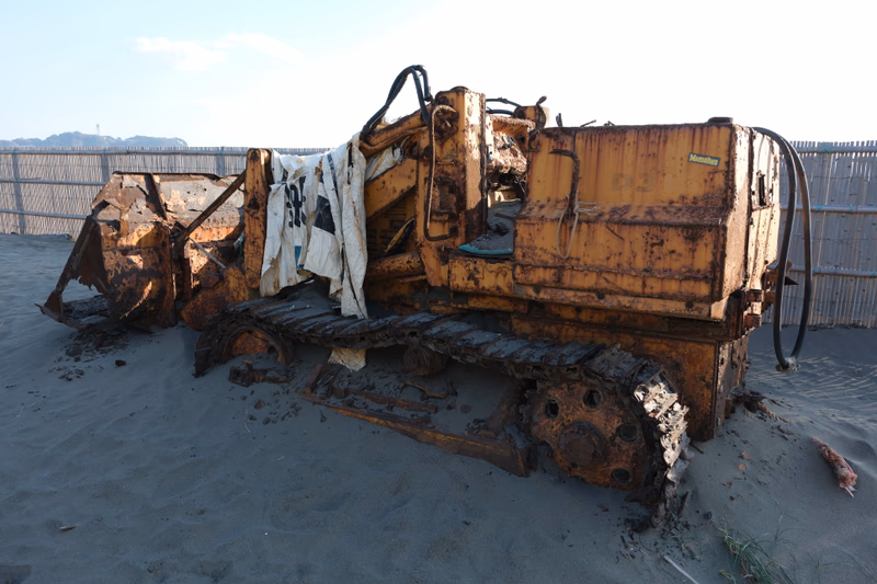 A rusty bulldozer sits on a sandy beach, covered in dust and debris, with a piece of cloth draped over it. The bulldozer is surrounded by a fence, and the scene is lit by the sun.