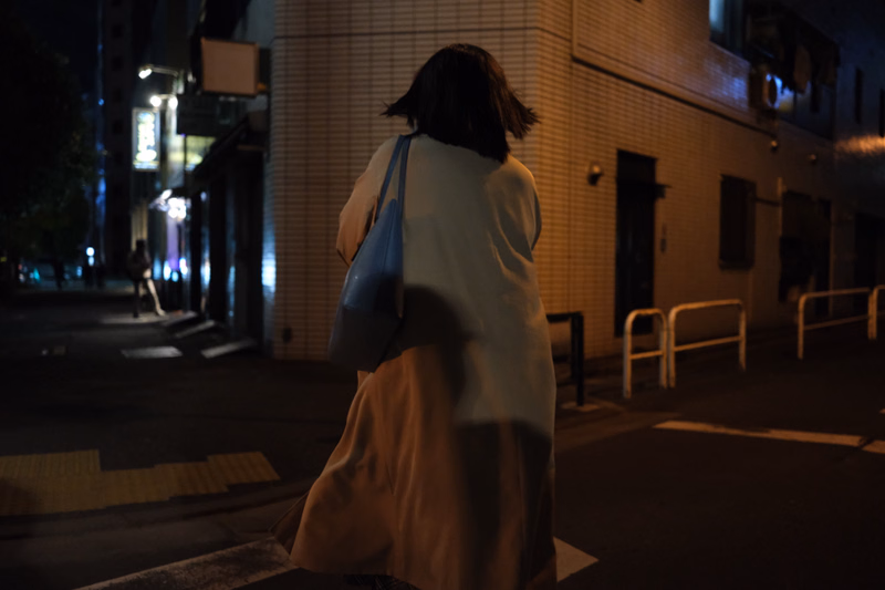 A person wearing a light-colored coat walks away from the camera on a dark city street at night, carrying a blue shoulder bag.
