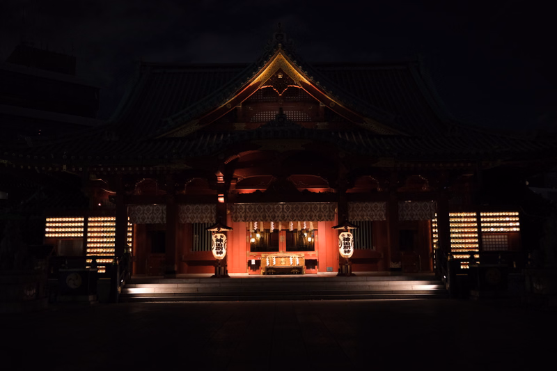 A large, traditional Japanese temple building illuminated at night, featuring a prominent tiled roof and red facade.