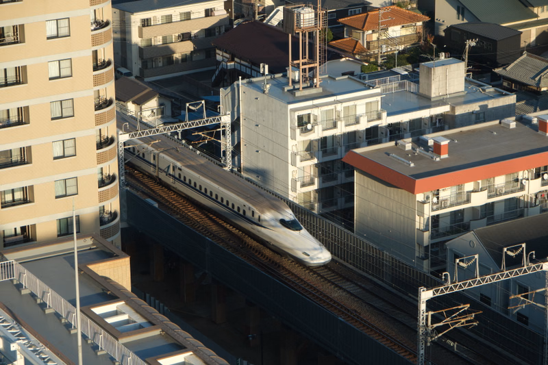 A modern train travels along elevated tracks through a dense urban area surrounded by multi-story buildings.