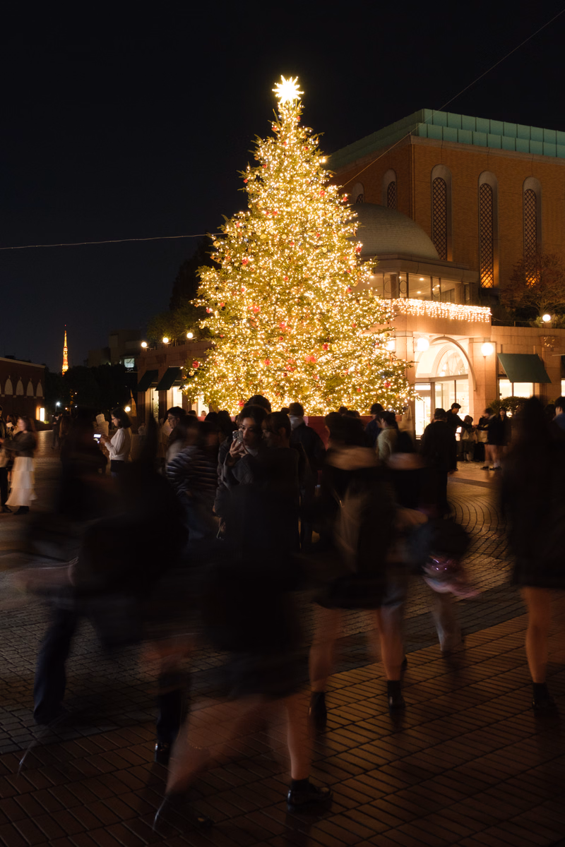 A brightly lit Christmas tree stands in a plaza surrounded by a blurred crowd of people at night.
