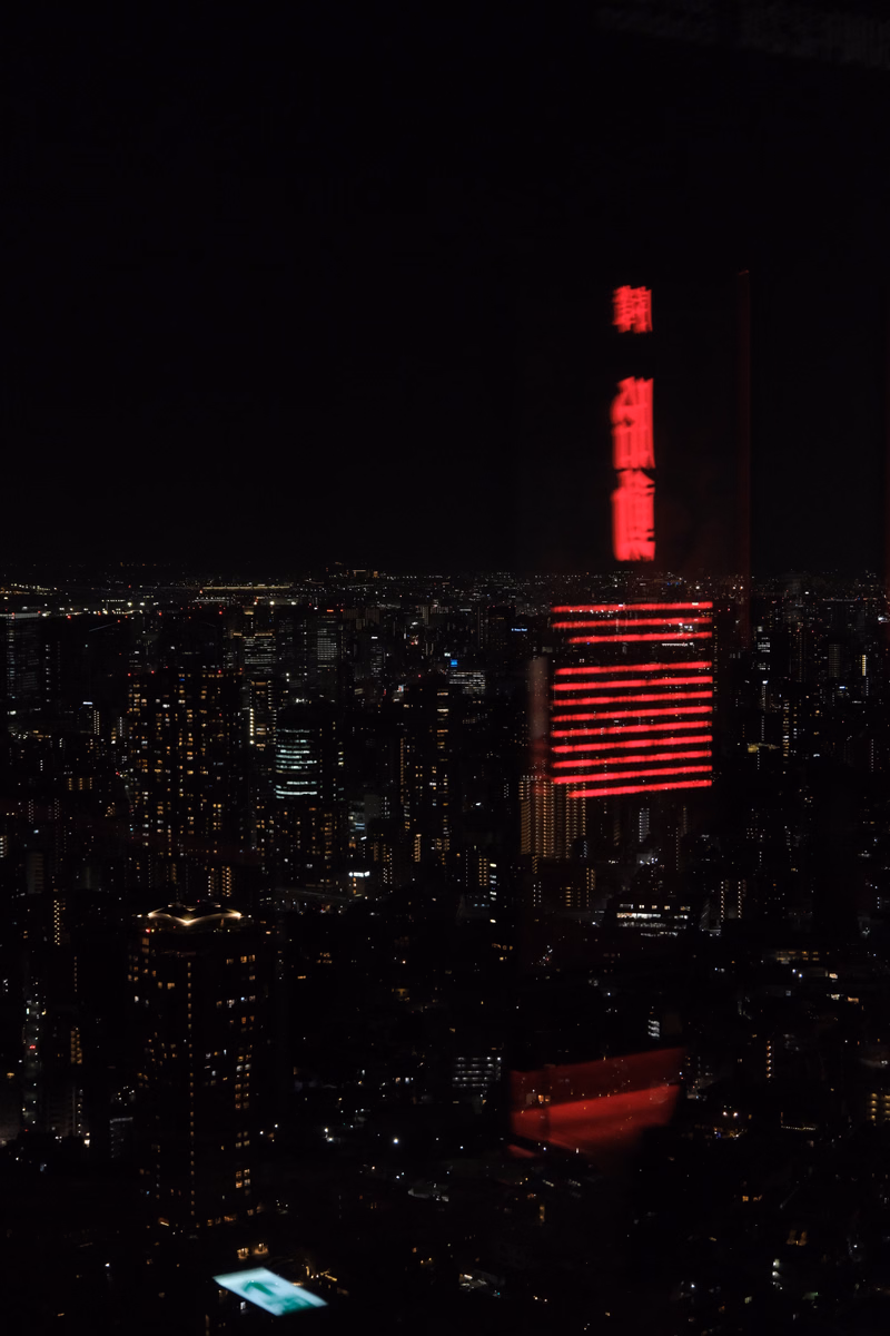 A nighttime cityscape view featuring a tall building with a bright red vertical billboard displaying Japanese characters.