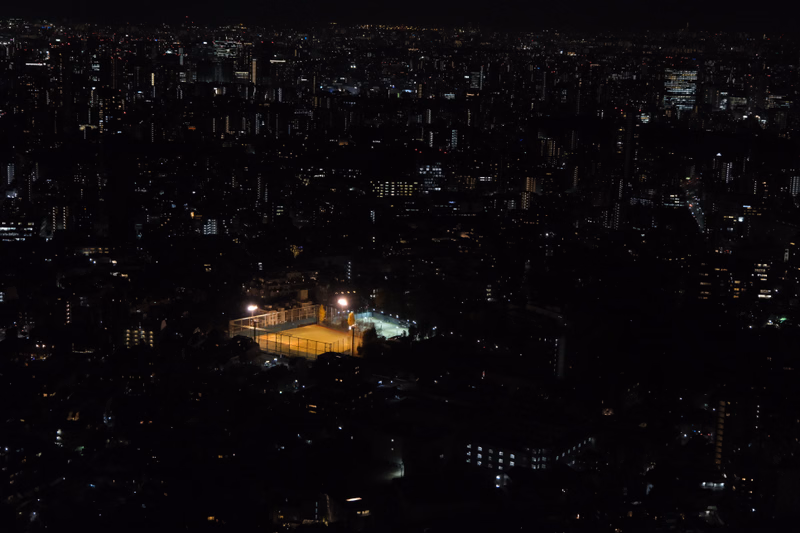 A nighttime aerial view of a sprawling city with numerous illuminated buildings and a brightly lit sports field in the foreground.