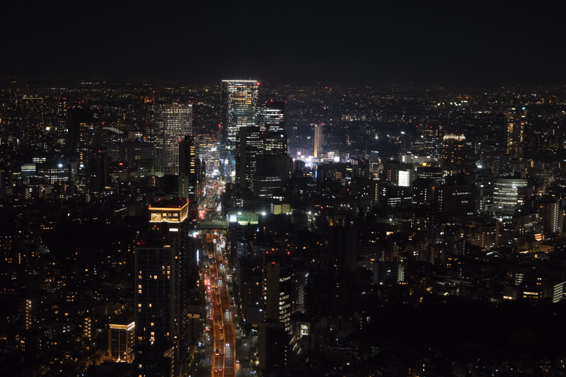 A wide nighttime cityscape view showing illuminated buildings and a central street running through the dense urban area.