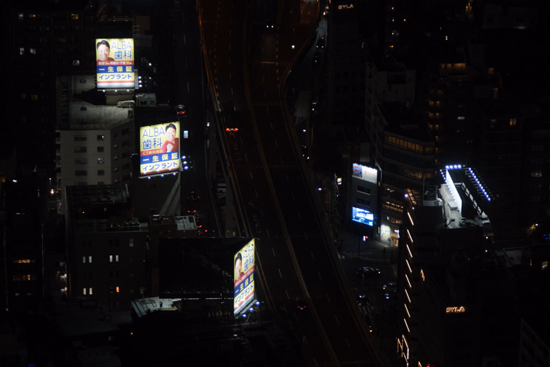 A nighttime cityscape view showing illuminated billboards, including one advertising "ALBA Dental Clinic," alongside elevated train tracks.