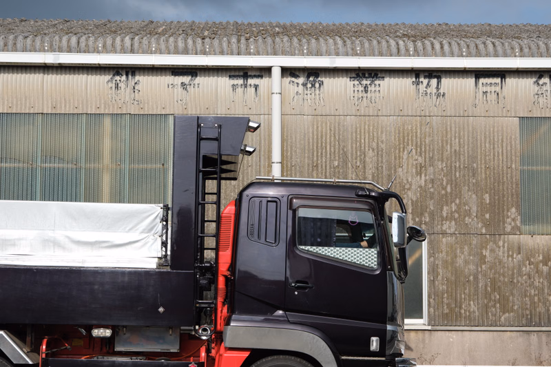 A dark purple flatbed truck is parked in front of a large, light-colored industrial building with corrugated siding.