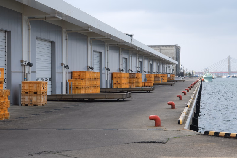 A long, modern warehouse building lines a concrete dock next to the water, with stacked wooden crates visible.