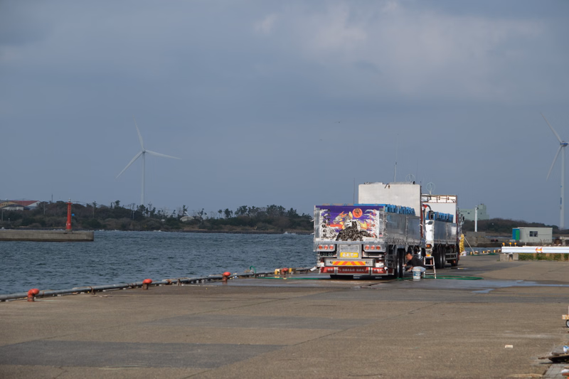 A large truck with a colorful cargo container is parked near a dock with water and several wind turbines visible in the background.