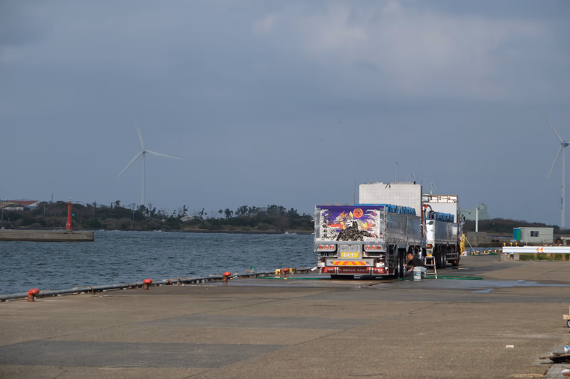 A truck is parked near a body of water with wind turbines in the background.