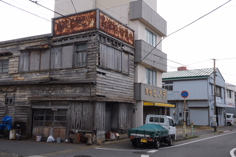 An old, weathered wooden building with a rusted sign sits next to a modern commercial building with a sign that reads "麺処ヒノタ".