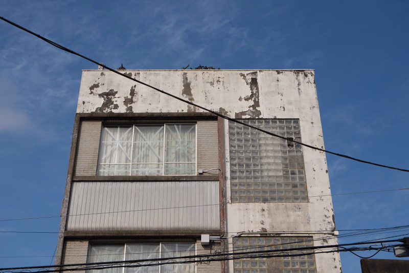 A weathered, multi-story concrete apartment building stands against a bright blue sky, with several overhead wires crossing the frame.