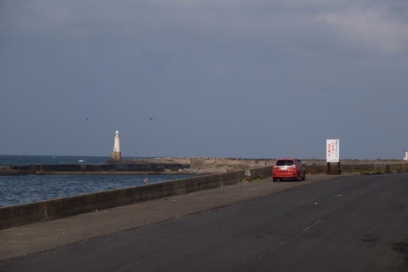 A white lighthouse stands near a seawall next to a paved road, with a red car parked nearby under a cloudy sky.