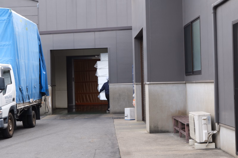 A white truck covered with a blue tarp is parked near the loading dock entrance of a modern gray building.