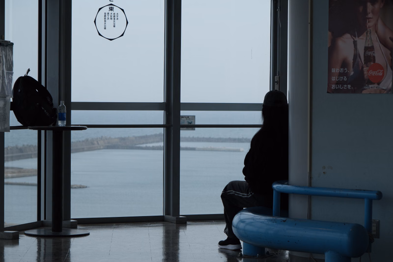 A person sits by a large window overlooking the ocean, with a Coca-Cola advertisement visible on the wall.