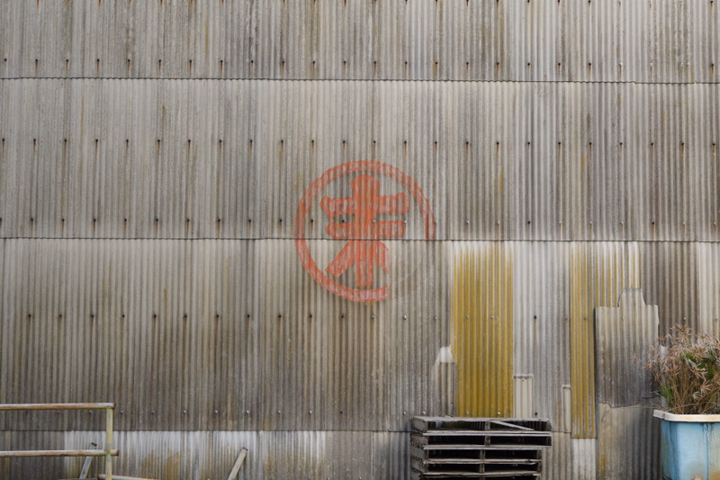 A large, weathered corrugated metal wall features a faded red circular logo or graffiti design.