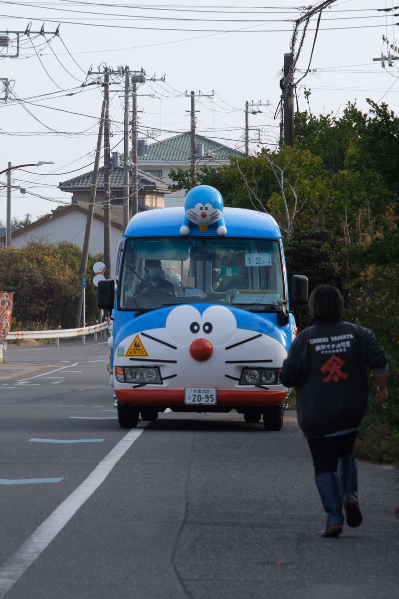 A blue bus decorated like a character is driving down a street, with a person walking in the foreground.