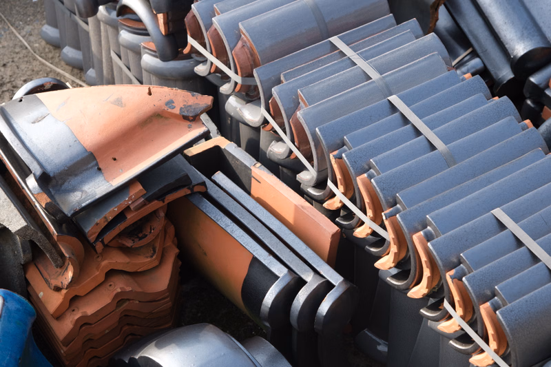 A stack of new grey interlocking roof tiles is next to a pile of reddish-brown clay roof tiles on the ground.