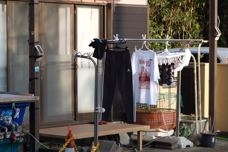 A clothes rack outside a beach hut displays black shorts and a white t-shirt that reads "EVA:02's P.M."