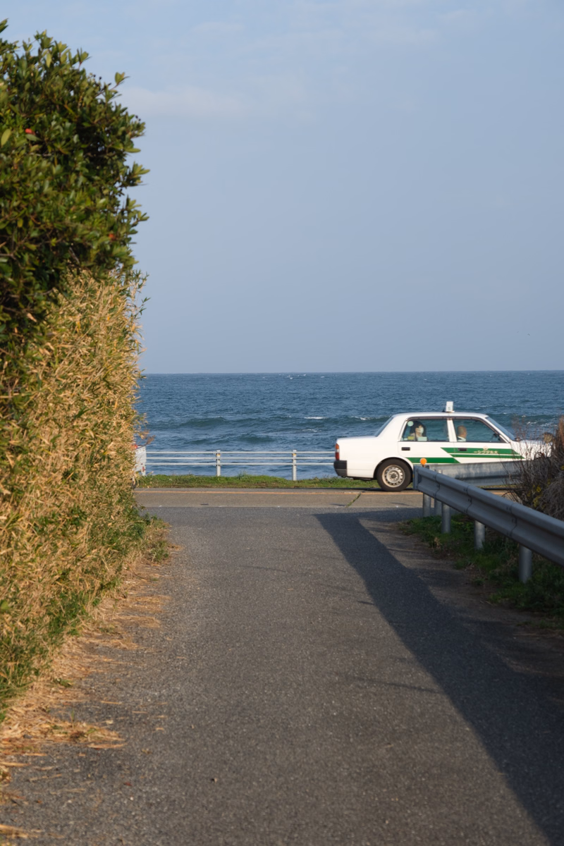 A white car is parked near a coastal road overlooking the sea under a clear sky, with trees lining the left side.