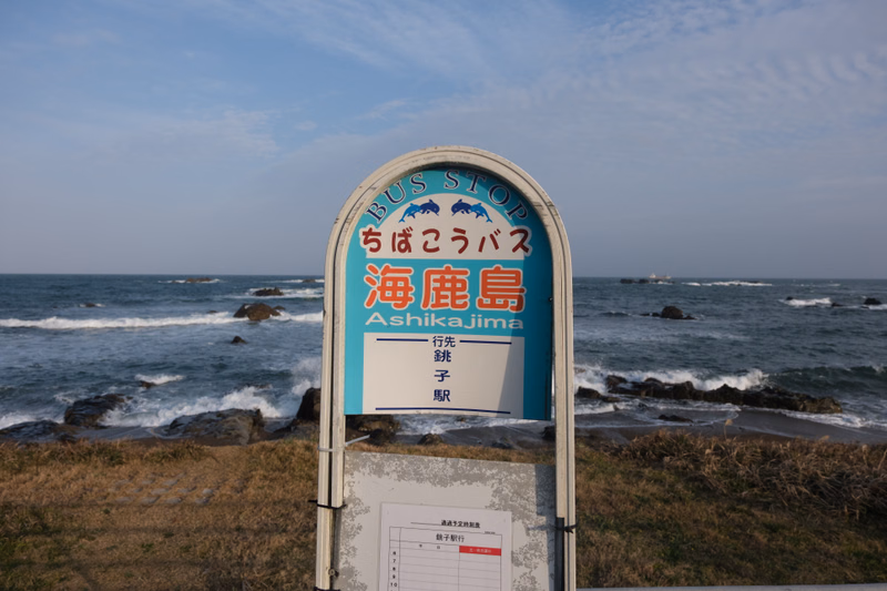 A bus stop sign for "ちばこうバス 海鹿島 Ashikajima" is visible near the ocean on a clear day.
